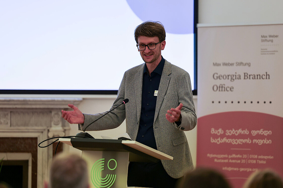 Ein Mann im Anzug steht an einem Pult mit dem Logo des Gothe-Instituts. Er hält die Arme geöffnet und spricht in Richtung des Publikums, von dem nur Hinterköpfe zu sehen sind. Hinter ihm steht ein Aufsteller auf dem "Georgia Branch Office" steht.