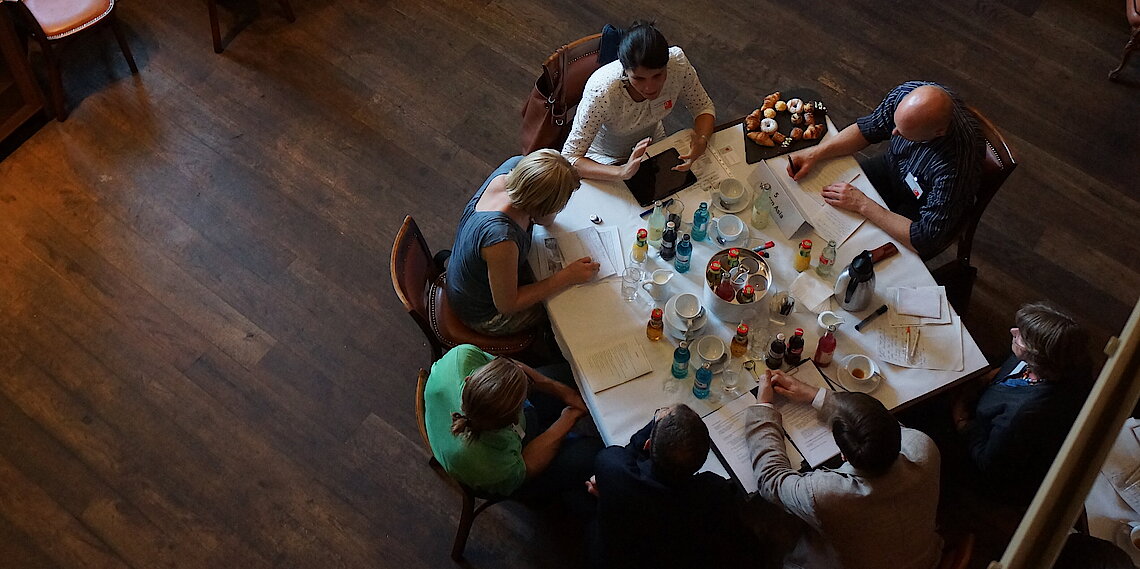 Vogelperspektive von sechs Personen, die an einem rechteckigen Tisch sitzen, der mit Papieren, Stiften, Flaschen und Gläsern bedeckt ist, in einem Raum mit Holzboden.