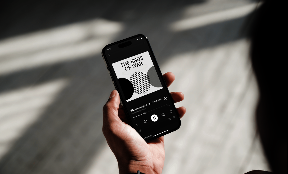 Eine Hand hält ein Smartphone, auf dessen Display der Podcast „The Ends of War“ zu sehen ist. Der Podcast gehört zur Themenreihe „Wissen entgrenzen“ der Max Weber Stiftung (MWS). Das Display zeigt das Titelbild des Podcasts mit grafischen Elementen in Schwarz, Weiß und Grau sowie den Wiedergabebildschirm mit Play-Button und Zeitangaben.