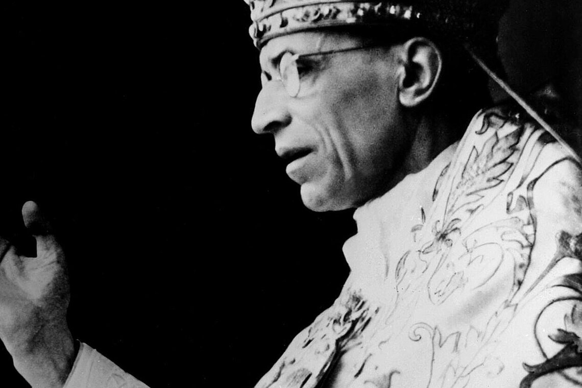 Schwarz-weiß-Fotografie einer Person in reich verzierter liturgischer Kleidung mit erhobener Hand.