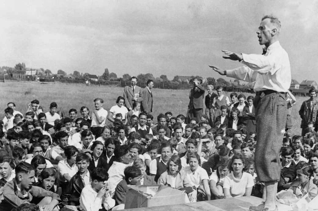 Schwarz-weißes Foto einer großen Menschenmenge im Freien. Im Vordergrund stehen und sitzen viele Jugendliche dicht gedrängt auf einer Wiese. Rechts im Bild steht ein erwachsener Mann. Er trägt ein helles Hemd und dunkle Hosen und spricht mit erhobenem Armen zu der Menge. Die Zuhörenden schauen aufmerksam zu ihm. Im Hintergrund sind offene Felder und vereinzelte Bäume zu sehen, der Himmel ist bewölkt. 